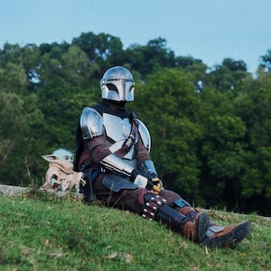 May include: A person in a Mandalorian costume sits on a grassy hill with a small Grogu figure. The costume features a silver helmet and armor, with brown and black clothing. The background includes lush green trees and a clear blue sky.