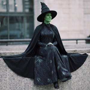Può includere: Una persona in costume da strega nero, con un cappello a punta alto e una lunga cappa. L'abito presenta un motivo a vortice con accenti verdi. La pelle della persona è dipinta di verde e siede su un davanzale di pietra.