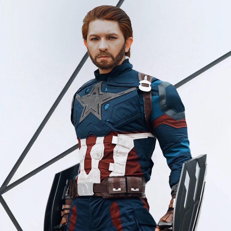May include: A person in a Captain America costume, featuring a blue and red suit with a white star on the chest. The costume includes a brown belt, a shield, and a matching helmet. The person has red hair and a beard.