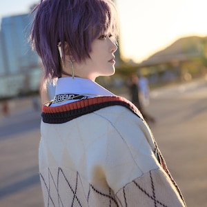 May include: Cream-colored argyle sweater with black diamond pattern, a white collar, and a red and black striped trim. The word "LEBEND" is printed on the collar. The person in the image has purple hair and is wearing long silver earrings.