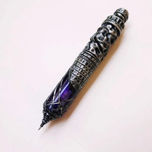 Op de afbeelding: Een decoratieve pen met een zilverkleurige, ingewikkeld gesneden buitenkant en een dieppaarse, diamantvormige loop. De pen heeft een puntige punt en een gedetailleerd, gestructureerd ontwerp. Het ontwerp van de pen doet denken aan fantasie- of historische artefacten.