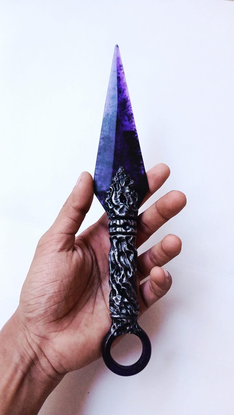 May include: A decorative kunai knife with a purple blade and a detailed, ornate handle. The handle is silver and black with a ring at the base. The blade is a deep purple color.