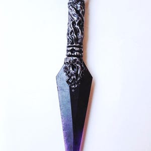 May include: A decorative kunai knife with a dark blade and a purple handle. The handle features intricate, textured detailing, and the blade transitions from black to purple. The knife is set against a white background.