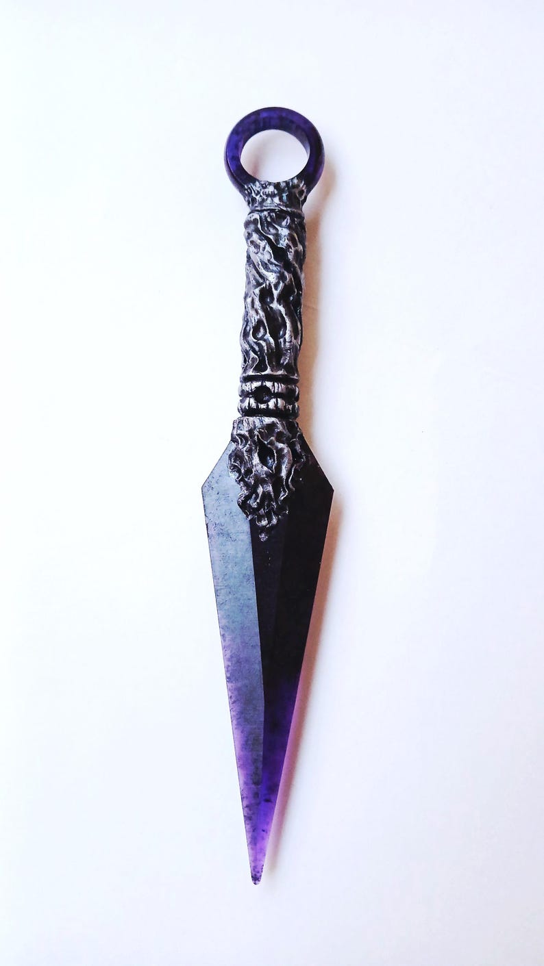 May include: A decorative kunai knife with a dark blade and a purple handle. The handle features intricate, textured detailing, and the blade transitions from black to purple. The knife is set against a white background.