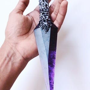 May include: A decorative dagger with a purple and clear crystal blade. The handle is silver with intricate carvings and a purple ring at the top. The dagger is held against a white background.
