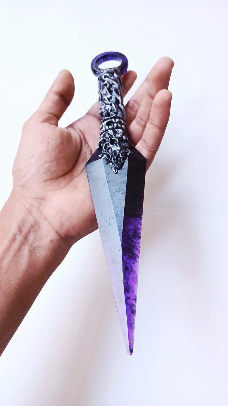 May include: A decorative dagger with a purple and clear crystal blade. The handle is silver with intricate carvings and a purple ring at the top. The dagger is held against a white background.