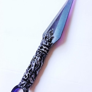 May include: A decorative kunai knife with a purple and silver color scheme. The blade is a gradient of purple and blue, while the handle features intricate, sculpted details. A circular ring is at the base of the handle.