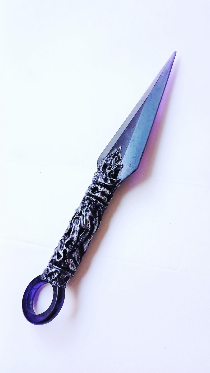May include: A decorative kunai knife with a purple and silver color scheme. The blade is a gradient of purple and blue, while the handle features intricate, sculpted details. A circular ring is at the base of the handle.