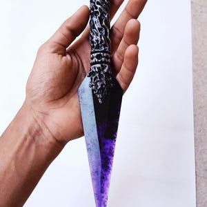 May include: A decorative kunai knife with a purple and black blade and a silver handle. The handle has intricate detailing and a circular purple ring at the top. The blade has a gradient effect, transitioning from black to purple.