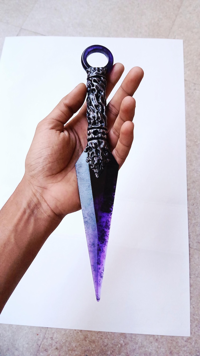 May include: A decorative kunai knife with a purple and black blade and a silver handle. The handle has intricate detailing and a circular purple ring at the top. The blade has a gradient effect, transitioning from black to purple.