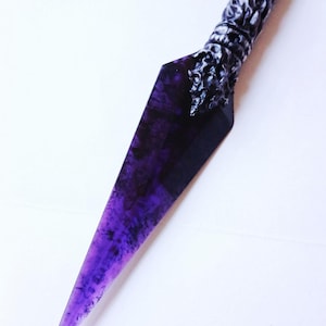 May include: A purple and black dagger with a textured handle and a ring at the end. The blade has a pointed tip and a marbled appearance. The handle has a detailed, ornate design.