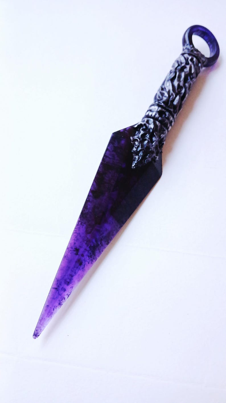 May include: A purple and black dagger with a textured handle and a ring at the end. The blade has a pointed tip and a marbled appearance. The handle has a detailed, ornate design.