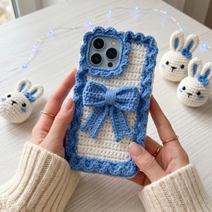 Puede incluir: Una funda de tel&eacute;fono de ganchillo blanca y azul con un lazo y bordes festoneados. La funda est&aacute; rodeada de peque&ntilde;as figuras de conejitos de ganchillo con lazos azules. La funda la sostiene una persona.