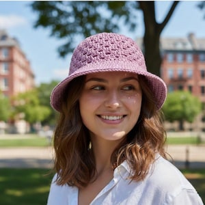 May include: A lavender crochet bucket hat with a wide brim. The hat has a textured, open weave design. The hat is worn by a person in a white button-down shirt, with a blurred background of trees and buildings.