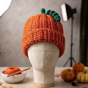 May include: A bright orange crocheted beanie hat with a ribbed texture and a folded brim. The top of the hat features a small, dark green crocheted stem and a curled vine detail, mimicking the appearance of a pumpkin.