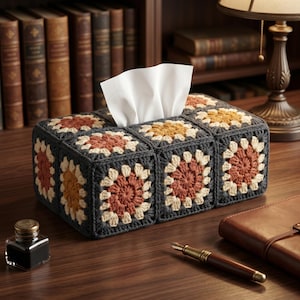 Op de afbeelding: Een handgemaakte tissueboxhoes met een granny square design. De hoes is donkergrijs met crème, oranje en gele bloempatronen. Witte tissues zijn zichtbaar aan de bovenkant. Op een houten bureau.