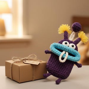 Puede incluir: Un monstruo de ganchillo morado hecho a mano con boca azul, detalles amarillos y dientes blancos est&aacute; junto a una caja de regalo marr&oacute;n atada con hilo. La caja de regalo tiene una etiqueta que dice "Handmade".