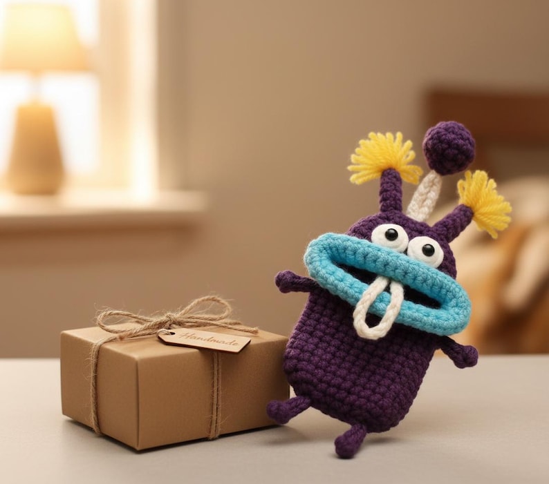 Puede incluir: Un monstruo de ganchillo morado hecho a mano con boca azul, detalles amarillos y dientes blancos est&aacute; junto a una caja de regalo marr&oacute;n atada con hilo. La caja de regalo tiene una etiqueta que dice "Handmade".