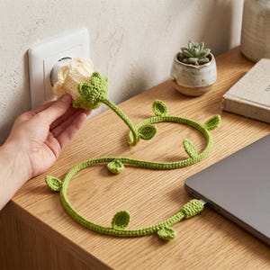 May include: A crocheted flower-shaped plug cover and cable wrap in cream and green. The cable, designed to look like a vine with leaves, is connected to a laptop. The flower is being plugged into a wall outlet. A small potted succulent and a book are on the wooden surface.
