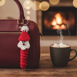 May include: A red crocheted Santa Claus keychain with a white beard and hat trim hangs from a burgundy handbag. A mug of hot chocolate with marshmallows steams in front of a fireplace, creating a cozy holiday scene.