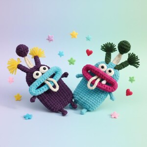 Puede incluir: Dos juguetes de monstruos de ganchillo hechos a mano en morado y azul, con caracter&iacute;sticas caprichosas. El monstruo morado tiene detalles amarillos, mientras que el monstruo azul tiene detalles verdes. Ambos tienen ojos grandes, bocas abiertas y lenguas blancas. Estrellas y corazones de ganchillo dispersos.