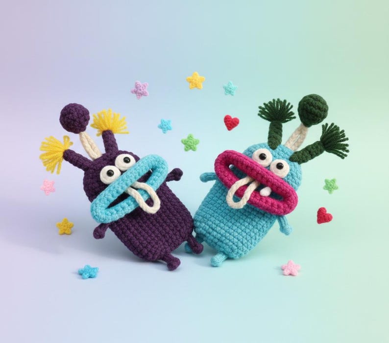 Puede incluir: Dos juguetes de monstruos de ganchillo hechos a mano en morado y azul, con caracter&iacute;sticas caprichosas. El monstruo morado tiene detalles amarillos, mientras que el monstruo azul tiene detalles verdes. Ambos tienen ojos grandes, bocas abiertas y lenguas blancas. Estrellas y corazones de ganchillo dispersos.