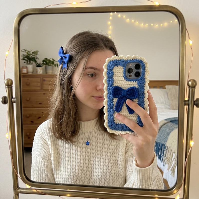 Puede incluir: Un lazo azul para el pelo, un collar con un lazo azul y una funda de tel&eacute;fono de ganchillo azul y blanco con un dise&ntilde;o de lazo se reflejan en un espejo. El espejo tiene un marco dorado.