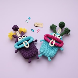 Könnte beinhalten: Zwei handgefertigte gehäkelte Monstertiere in Lila und Blau, mit großen Augen und farbenfrohen Akzenten. Das lila Monster hat gelbe und lila Akzente, während das blaue Monster grüne und rosa Akzente hat. Auf hellviolettem Hintergrund.