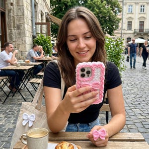 Puede incluir: Una funda de tel&eacute;fono rosa y blanca de ganchillo con un dise&ntilde;o de lazo. La funda tiene un borde festoneado texturizado y un lazo en la parte delantera. Tambi&eacute;n se ve un anillo de lazo de ganchillo a juego. Los art&iacute;culos est&aacute;n hechos a mano.