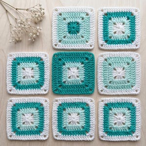 Puede incluir: Nueve posavasos cuadrados de ganchillo en blanco, turquesa y azul claro. Cada posavasos presenta un diseño cuadrado central, bordeado por un borde turquesa y un borde exterior blanco. Los posavasos están dispuestos sobre una superficie de madera clara.