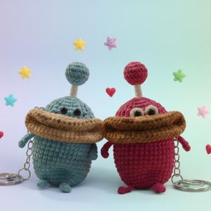 May include: Two handmade crochet keychains, one teal and one red, each with a unique alien-like design. They feature brown scarves, googly eyes, and silver key rings. The background includes colorful stars and hearts.