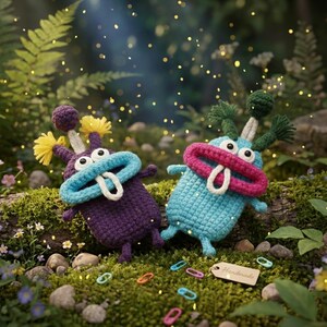 Puede incluir: Dos juguetes de monstruos hechos a mano con ganchillo en un entorno forestal. Uno es morado con detalles amarillos, el otro es azul con detalles rosas. Ambos tienen bocas grandes, lenguas blancas y ojos grandes. Los juguetes est&aacute;n rodeados de musgo, flores y clips de colores.