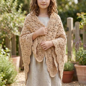 May include: A beige crochet shawl with a floral design. The shawl features a scalloped border and is worn over a light-coloured dress. The image is taken outside, with a wooden fence and plants in the background.
