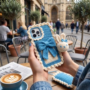 Puede incluir: Funda de tel&eacute;fono de ganchillo azul y crema con un dije de conejito. La funda presenta un lazo grande y bordes festoneados. Tambi&eacute;n se ve una peque&ntilde;a funda a juego. Escena borrosa de un caf&eacute; al aire libre.