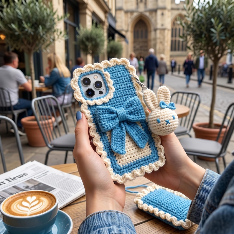 Puede incluir: Funda de tel&eacute;fono de ganchillo azul y crema con un dije de conejito. La funda presenta un lazo grande y bordes festoneados. Tambi&eacute;n se ve una peque&ntilde;a funda a juego. Escena borrosa de un caf&eacute; al aire libre.