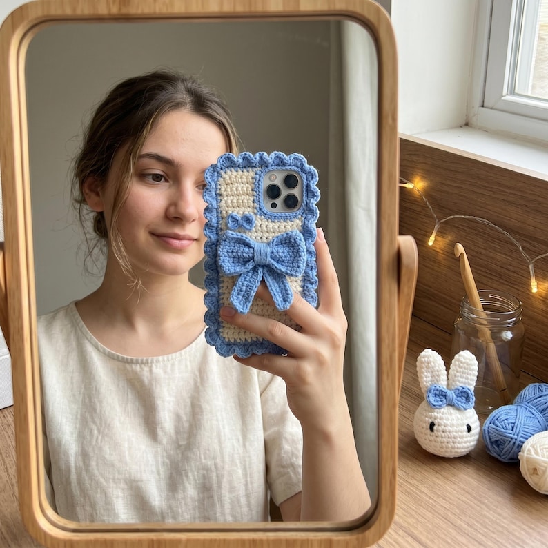 Puede incluir: Una funda de tel&eacute;fono de ganchillo con un lazo y ribete azul claro. La funda es de color crema con un borde festoneado azul. La funda del tel&eacute;fono se sostiene frente a un espejo. Un conejito de ganchillo y ovillos de lana son visibles en el fondo.