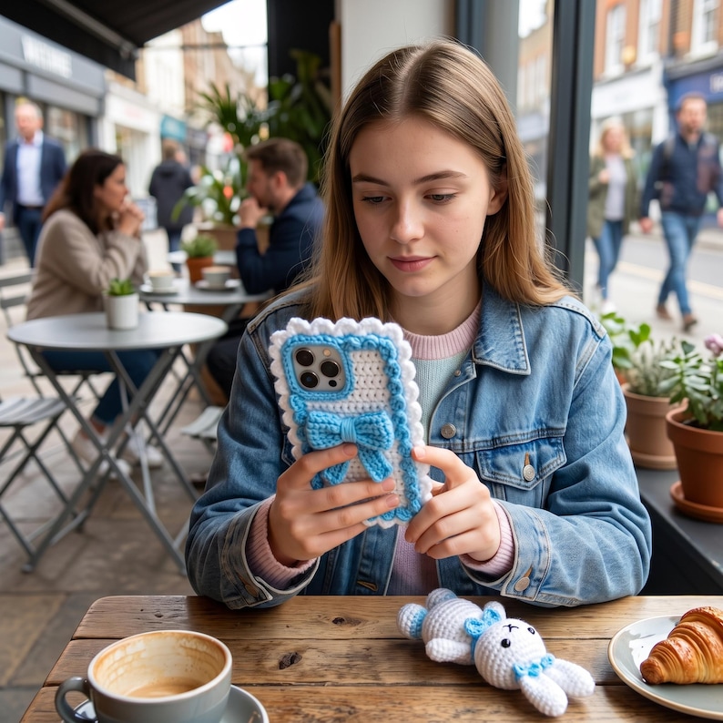 Puede incluir: Una funda de tel&eacute;fono de ganchillo blanca y azul con un detalle de lazo. La funda tiene un borde festoneado y la sostiene una persona. Un peque&ntilde;o conejito de ganchillo blanco y azul est&aacute; sobre la mesa de madera. Una taza de caf&eacute; y un cruas&aacute;n tambi&eacute;n est&aacute;n sobre la mesa.