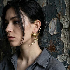 Op de afbeelding: Een goudkleurige ear cuff en oorbelset met een langwerpige, gebogen metalen structuur die rond het bovenste oorkraakbeen wikkelt en verbonden is met een bungelende, abstracte, puntige metalen hanger aan de oorlel.