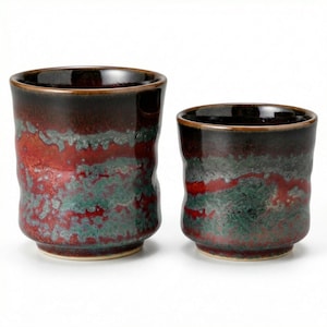 Op de afbeelding: Twee keramische bekers met een getextureerd oppervlak. De bekers hebben een donkerbruine rand en bodem, met een rood en groen gevlekt patroon in het midden. De bekers zijn van verschillende grootte.