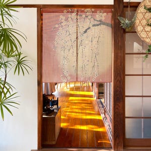Könnte beinhalten: Ein japanischer Vorhang, oder Noren, hängt in einem Türrahmen. Der Vorhang ist ein zartes Rosa mit einem floralen Design aus weißen Blüten und dunklen Ästen. Der Türrahmen führt in einen Raum mit Holzboden.
