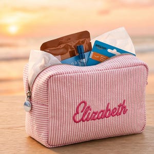May include: A rectangular pink and white striped zippered pouch featuring the name Elizabeth embroidered in dark pink script on the front. The pouch is filled with white tissues, a small clear spray bottle, a brown packet, and a blue Amazon gift card.