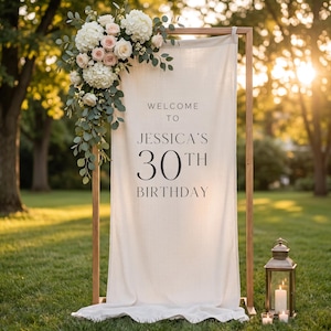 May include: A vertical cream-coloured fabric banner suspended from a wooden frame, featuring the black text WELCOME TO JESSICA'S 30TH BIRTHDAY. The top left corner is adorned with a floral arrangement of white hydrangeas, pale pink roses, and green eucalyptus leaves.