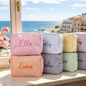 Könnte beinhalten: Eine Sammlung rechteckiger Reißverschlusstaschen mit vertikalem Nadelstreifenmuster in verschiedenen Pastellfarben. Jede Tasche ist mit einem Namen in kursiver Schreibschrift bestickt, darunter Ella, Luna, Lily und weitere sichtbare Namen.