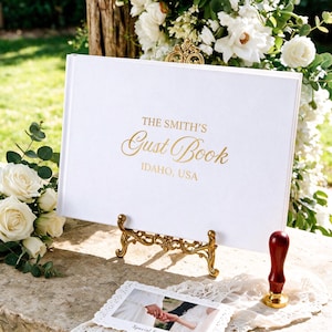 Puede incluir: Un libro de visitas blanco con texto en lámina dorada que dice THE SMITH'S Guest Book IDAHO, USA, exhibido en un soporte de metal dorado ornamentado. Un sello de cera rojo y una pequeña fotografía con borde de encaje están en primer plano.