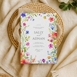 Pode incluir: Um convite de casamento com os nomes "SALLY" e "ADNAN" rodeados por uma borda floral em aquarela. O design inclui flores rosa, azuis e vermelhas. O convite também mostra a data, hora e local da cerimônia.