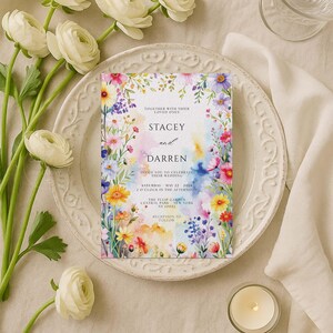 May include: A floral wedding invitation featuring the names "Stacey and Darren" with watercolor accents. The invitation is surrounded by colorful flowers and greenery. The text includes the date, time, and location of the wedding.