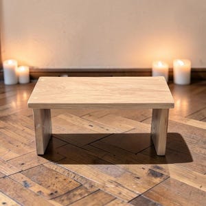 Peut inclure: Un petit tabouret rectangulaire en bois avec une finition naturelle couleur bois clair. Le tabouret présente une surface supérieure plate soutenue par deux panneaux latéraux verticaux, offrant un design simple et fonctionnel.