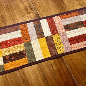 May include: A rectangular quilted table runner with a patchwork design. The runner features various autumn-themed fabrics in shades of yellow, orange, red, brown, and cream. The edges are finished with a dark brown border, creating a warm, inviting aesthetic.