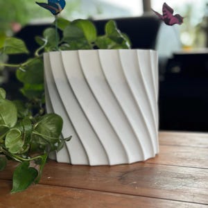 Op de afbeelding: Een witte, cilindervormige bloempot met een spiraalvormig ontwerp. De pot is gemaakt van een stevig materiaal en staat op een houten oppervlak. Groene bladplanten zijn zichtbaar in de pot. Decoratieve vlinders zijn op de achtergrond.