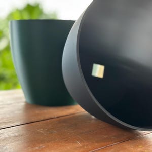May include: Two cylindrical planters, one dark green and the other black, are positioned on a wooden surface. The black planter is tilted, revealing its interior. The background is blurred, suggesting an outdoor setting.